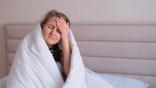 Une femme se sent mal à la maison assise dans son lit sous des couvertures blanches et fronçant les sourcils en touchant sa tête