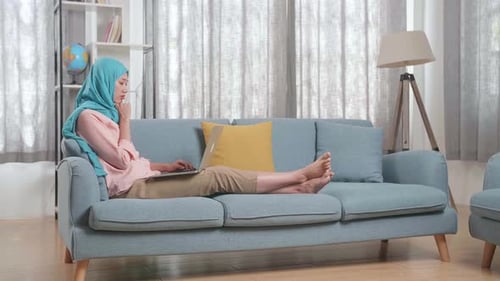 Uma mulher muçulmana se apoiando em um sofá e pensando em algo enquanto trabalha em um laptop em casa