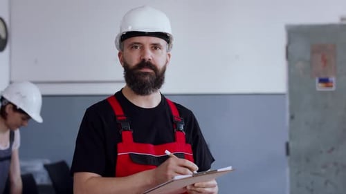 Mature engineer in hardhat writing on clipboard in bright industrial office