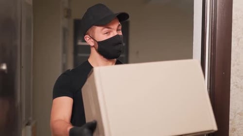 Young man safely delivers cardboard box to home in protective gloves and mask in black uniform