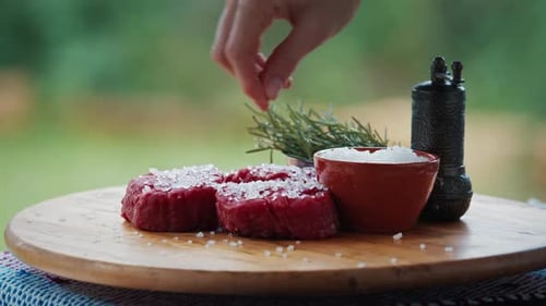 BBQ in the garden. Woman seasons filet mignon resting on the table with pepper and rosemary.