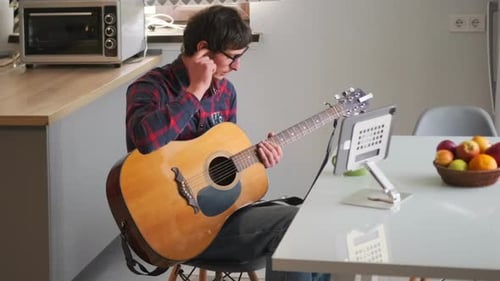 Jeune homme concentré dans des lunettes jouant du cou de guitare et étudiant depuis une tablette à la maison en gros plan