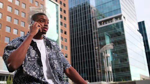 An African American Man Sits on the Street and Talks on His Smartphone in a Business District