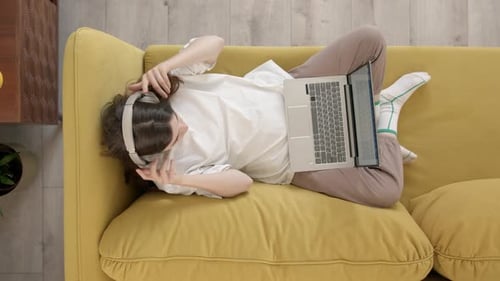 Top Down View of Young Woman in White Tshirt Lies on Yellow Sofa with Laptop on Her Lap Puts on