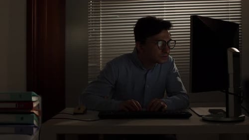 Young Man Sitting at His Office Desk in the Dark Removing His Eyeglasses While Concentrating on the