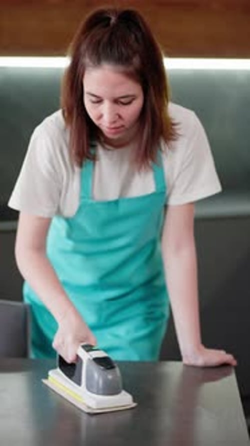 Vertical Video Portrait of a Brunette Cleaning Lady in a Blue Apron Who Polishes a Glossy Table
