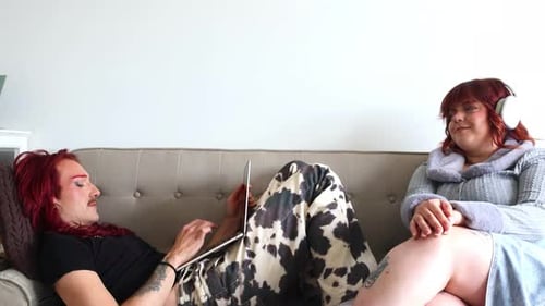 Non-binary person using laptop next to chubby friend wearing headphones