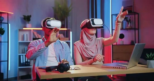 Man and woman in hijab working on imaginary screen in augmented reality goggles in home office