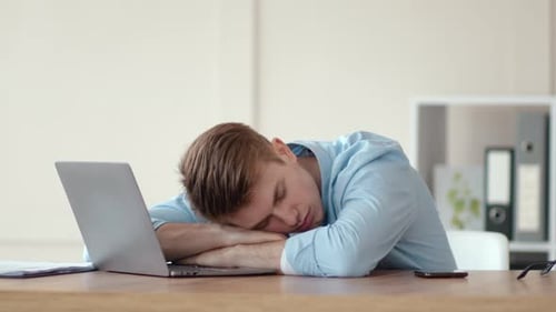 Müder junger Mann schläft am Arbeitsplatz auf dem Laptop