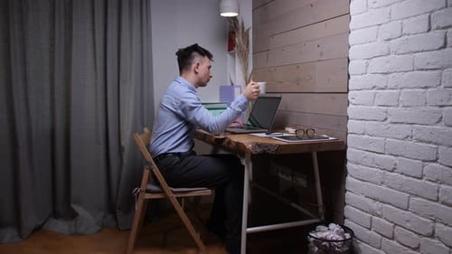 Young Man Telecommuting at His Home Office Desk Holding a Cup of Coffee While Concentrating on Work