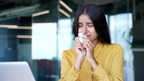 Close-up of sick young businesswoman blowing her nose with a handkerchief in modern office.