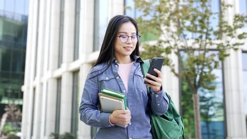 Smiling asian female student using phone walking on campus space near university building. A woman