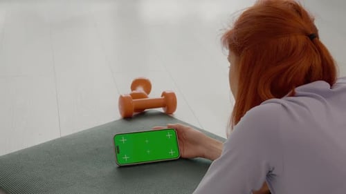 Woman relaxes on gym mat using smartphone with green screen for workout
