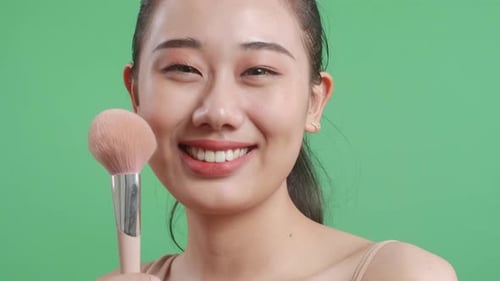 Close Up Of Beautiful Asian Woman Rouging Her Cheeks And Holding Makeup Brush On Green Screen