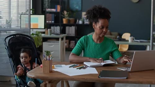 Woman Working on Laptop at Office Desk next to Little Son in Stroller