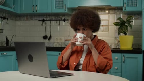 Woman Using Laptop and Drinking Coffee in Kitchen