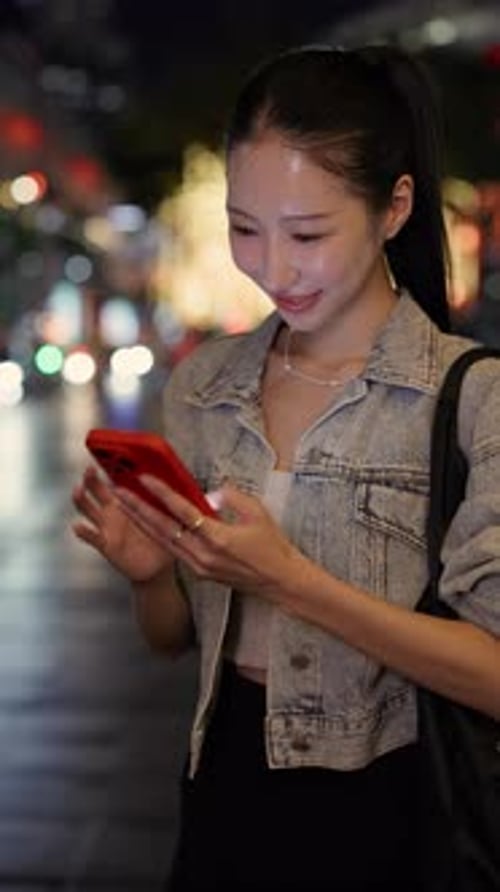 Portrait of Happy Asian Woman Texts on Smartphone and Stands in Night Downtown