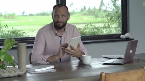 Man Uses Tablet at Table Indoors near Window