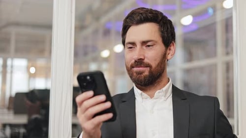 Businessman in Suit Smiles at Camera While Using Smartphone in Modern Office