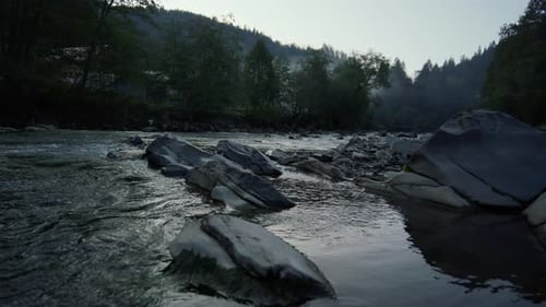 Abundant mountain river flows through forest trees over stone boulders at sunset