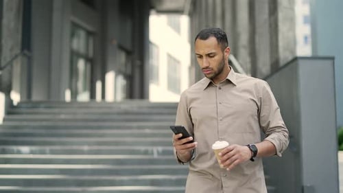 A young adult man is looking at a smartphone while walking down the street near an office building.