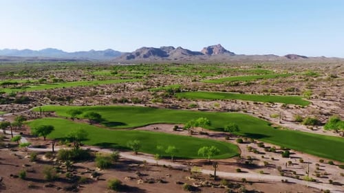 Wide aerial view of a desert golf course surrounded by rugged terrain and mountain ranges under clea