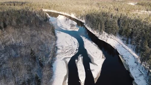 Aerial view of a winding river through a dense snow-covered forest in winter, with a curving road ru