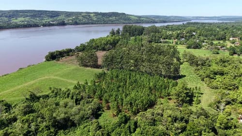 Wide River Bordering Green Farmland And Dense Forest Under Sunlight