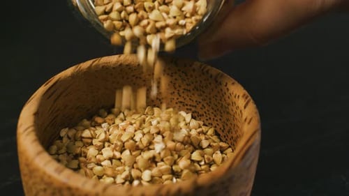 Pouring Raw Whole Grain Green Buckwheat Seeds From Glass Jar Into a Wooden Cup