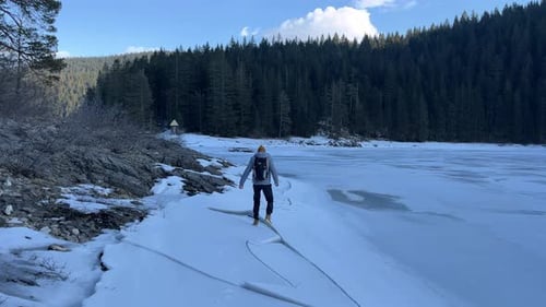 A Tourist with a Backpack Walks on Cracked Ice on a Frozen Forest Lake in Winter