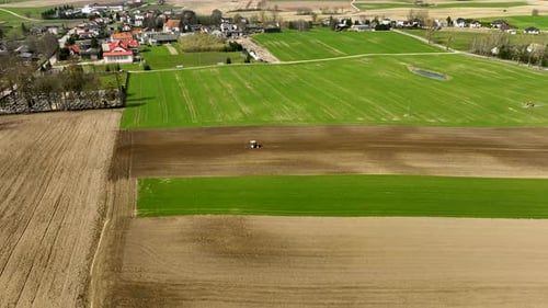 Aerial birds eye of plowing tractor on farm field near small village at sunny day in spring season.