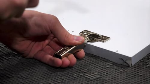 Close-up shot of a hand using a hammer to install a metal hinge on a white wooden panel, set on a
