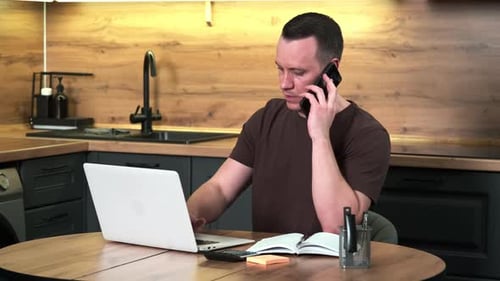 A Male Freelancer Works Sitting at the Kitchen Table Using a Phone and a Laptop