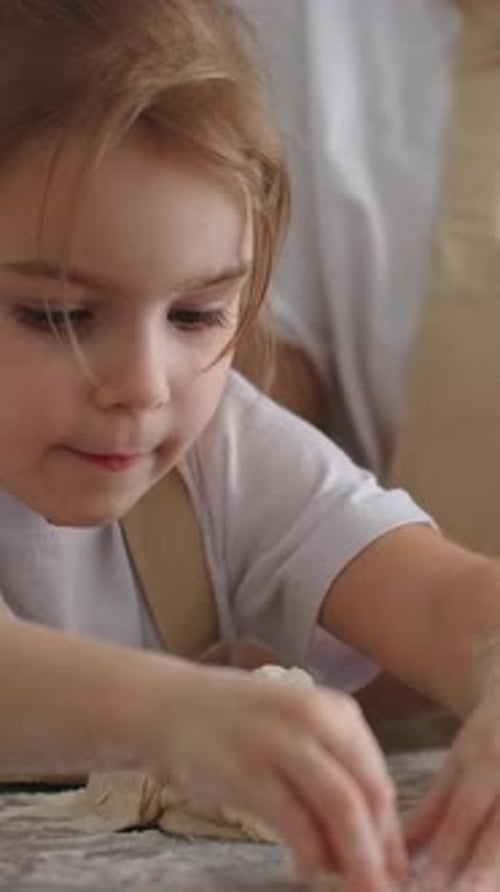 Kleines Kind knetet weichen Teig mit kleinen, mit Mehl bedeckten Händen neben einem Erwachsenen in einem gemütlichen Zuhause
