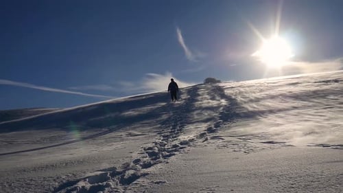 Man Walking in the Snow on a Mountain Plain covered with Powdered Snow Flowing in the Wind under a L