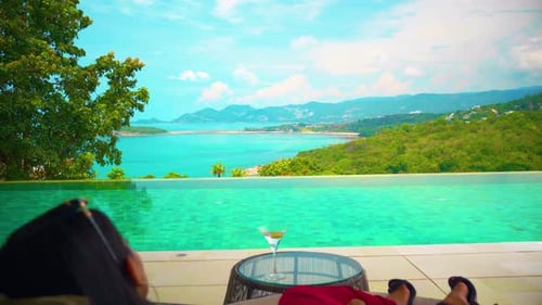 a woman with a sexy evening dress on a trip sitting on a sofa by the pool drinking and looking at th