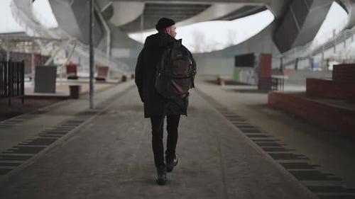 Young Man in Black Clothes Carrying Backpack and Walking in Urban Outskirts Back View of Hipster