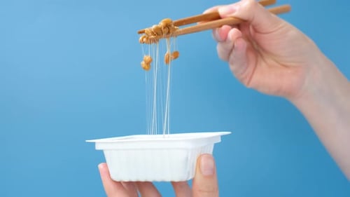Female hand holding Japanese natto beans with wooden chopsticks on a blue background, side view.