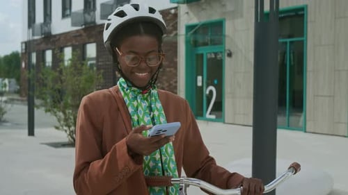 Smiling Lady in Helmet and Phone in Hand Rolling Bicycle to Office Building
