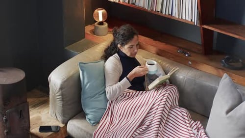 Biracial woman lying on sofa under blanket reading book and drinking coffee at home, slow motion