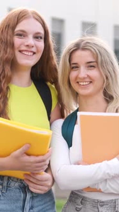 Two Smiling Female Students Holding Books on a Lively School Campus