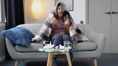 Young Man Suffering From a Common Cold and Flu or Allergy Sit at Home Wrapped in Blanket While He
