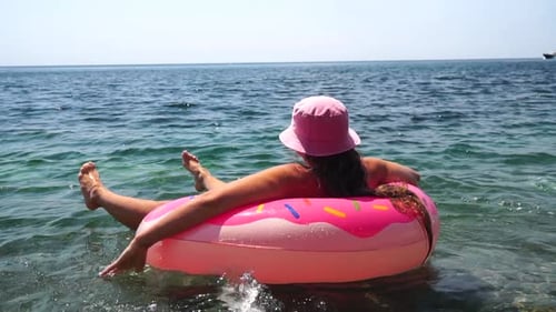 Férias de verão: Mulher engraçada de chapéu flutua em um colchão de donut inflável rosa, um anel de natação de brinquedo aquático