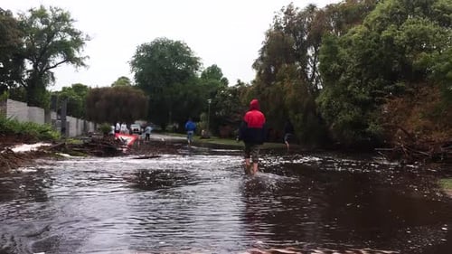 People walking on flooded street with broken down road sign amongst debris and car with hazards in b