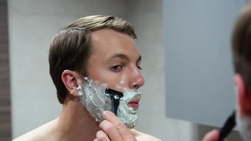 Man Shaves Face with Razor in Bathroom Mirror