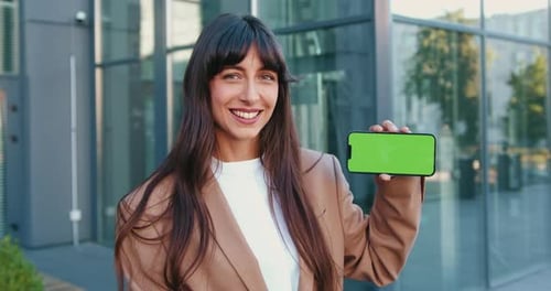 Smiling young business woman with long hair pointing at smartphone with green screen. Chroma key pho