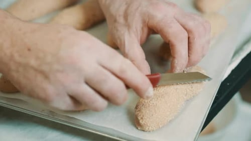 Bakery chef scoring bread rolls on baking sheet with knife, preparing sesame bread rolls for the ove