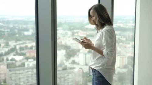 Woman Uses Tablet Standing by Apartment Window