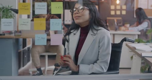 Creative Young Businesswoman in Wheelchair Working with Sticky Notes on Glassboard in Office Focused