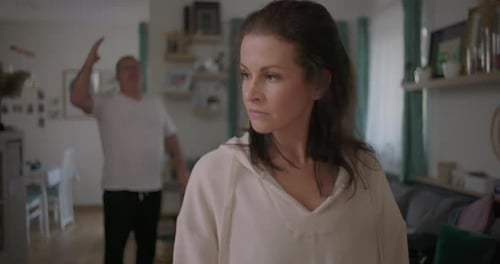 Woman standing with downcast expression in foreground during argument, man gesturing in frustration